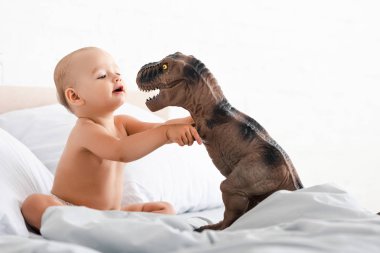 Küçük çocuk beyaz battaniye ile yatakta oturan ve iki eliyle oyuncak dinozor tutarak