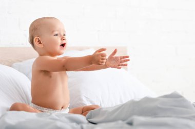 Küçük çocuk beyaz battaniye ile yatakta oturan ve ileri ellerini yükselterek