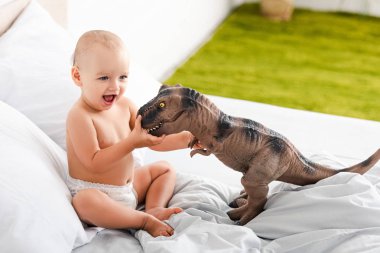 Cute little child smiling and playing with brown toy dinosaur on white bed clipart