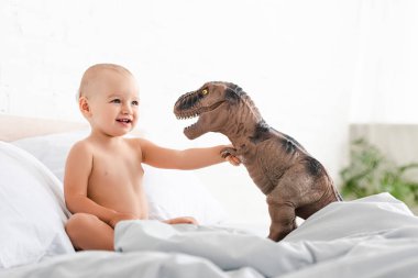 Cute little child sitting on bed, smiling and holding brown toy dinosaur  clipart