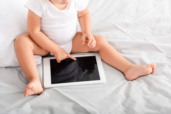 Cropped view of little child in white clothes sitting on bed and pointing with finger at digital tablet