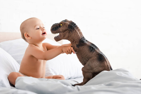 Little child sitting on bed with white blanket and holding toy dinosaur with both hands