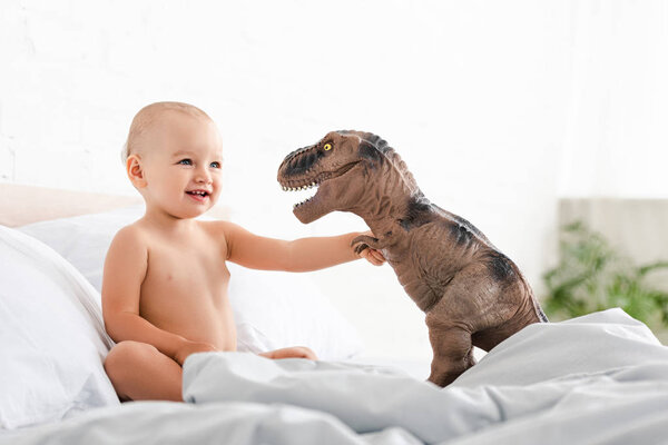 Cute little child sitting on bed, smiling and holding brown toy dinosaur 