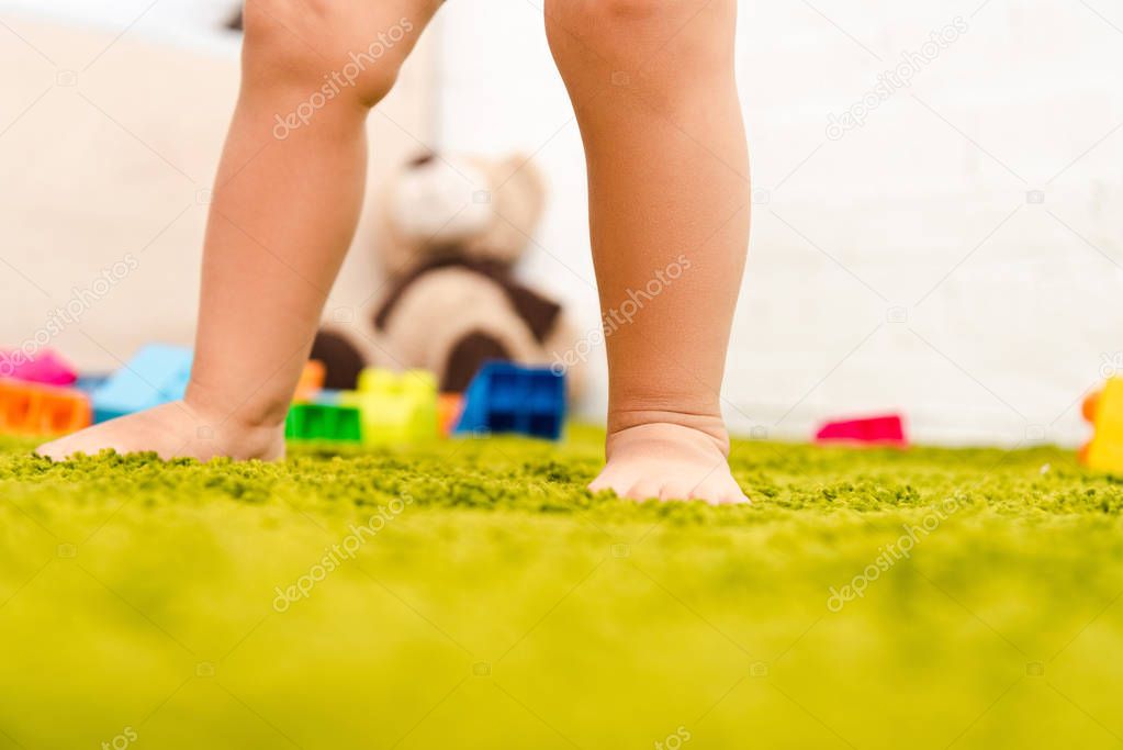 Cropped view of barefoot child standing on green floor among colorful toys