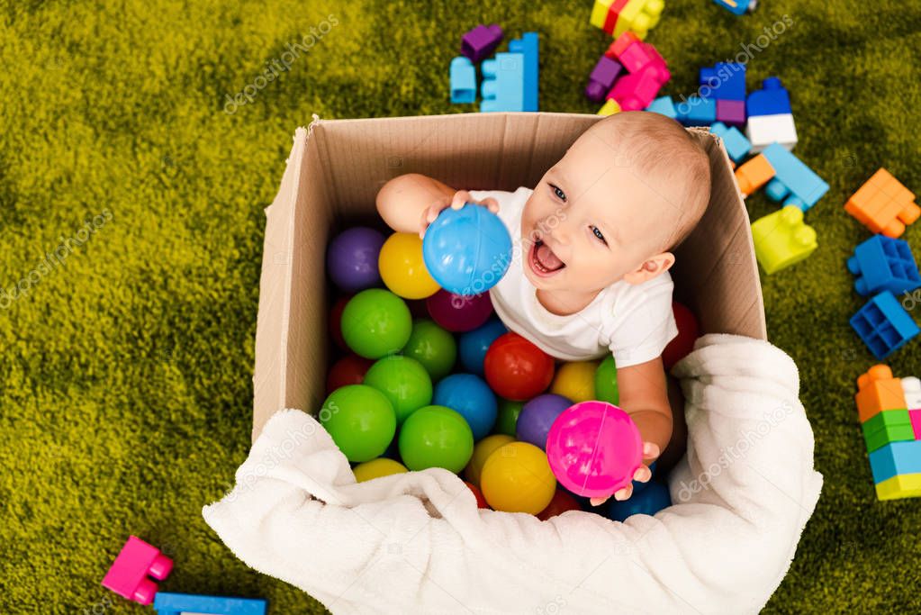 Happy little child sitting in cardboard box and playing with colorful balls