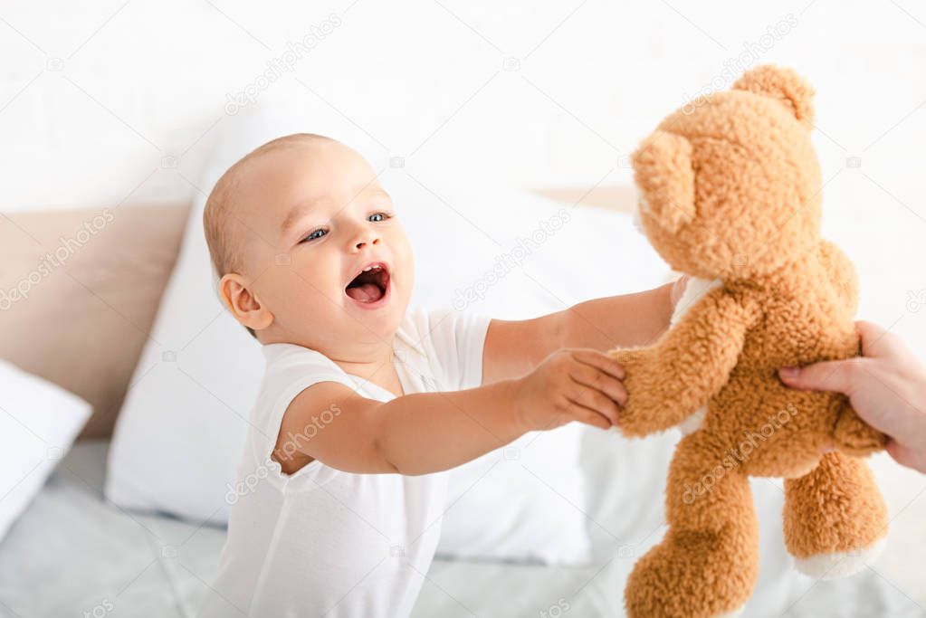 Cute little child in white clothes opening mouth, smiling and raising hands to brown plush bear 