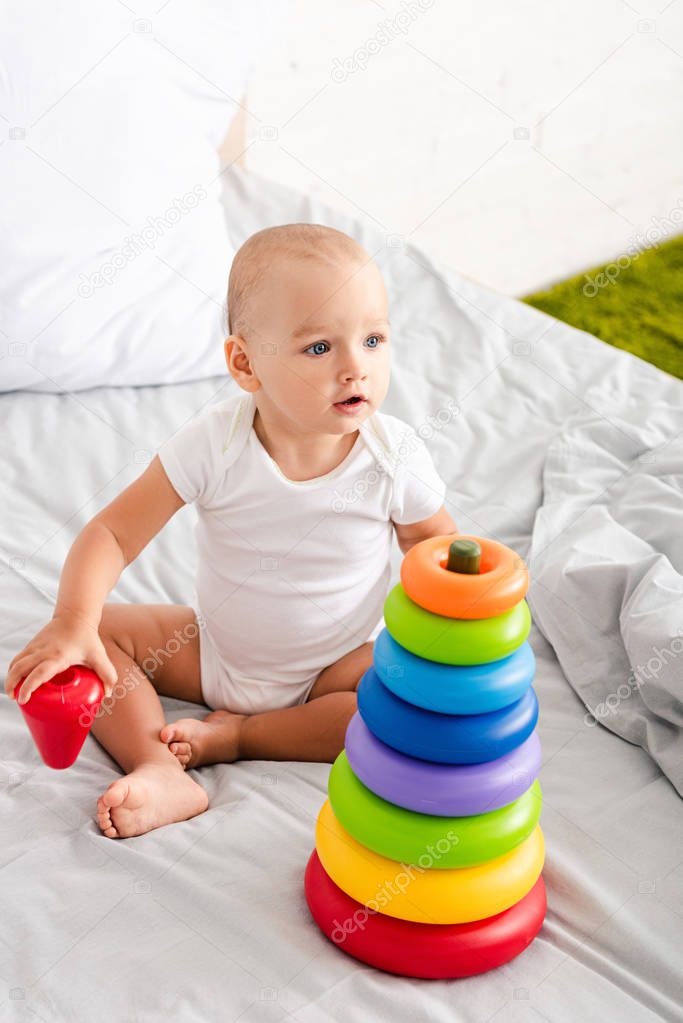 Funny barefoot child in white clothes playing with toy pyramid on bed