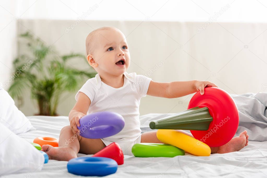 Cute little child in white clothes sitting on bed with colorful toy pyramid and smiling