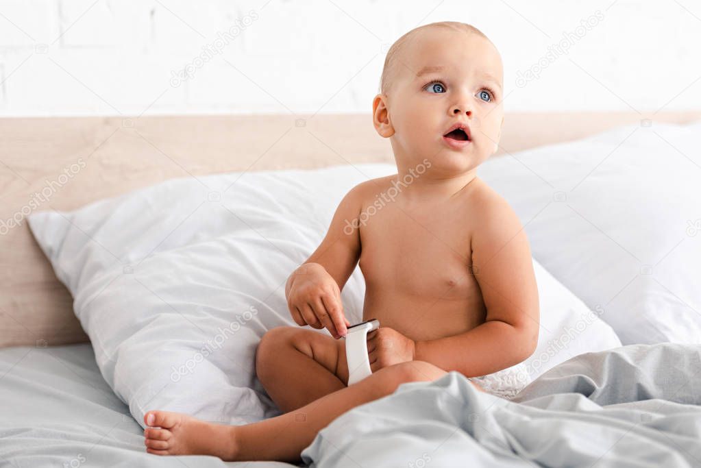 Cute baby sitting on white bed and holding smartwatches in hands