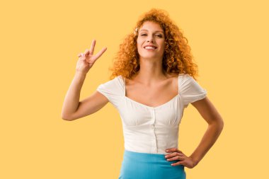 smiling woman with red hair showing victory symbol isolated on yellow clipart