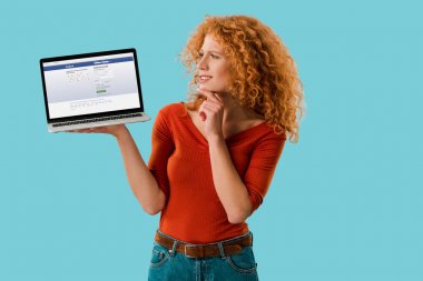 Kiev, Ukrayna - 16 Temmuz 2019: pensive redhead kadın web sitesi ile dizüstü facebook tutan, mavi izole 