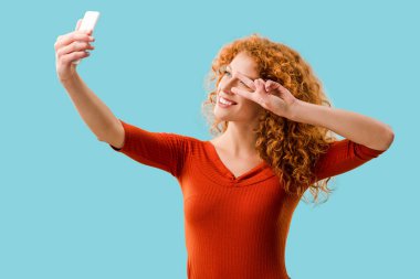 barış sembolü ile kıvırcık kızıl saçlı kadın mavi izole akıllı telefonda selfie alarak