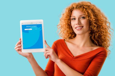 KYIV, UKRAINE - JULY 16, 2019: smiling redhead woman holding digital tablet with twitter app, isolated on blue  clipart