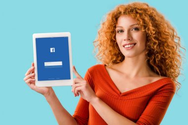 Kiev, Ukrayna - 16 Temmuz 2019: facebook uygulaması ile dijital tablet tutan gülümseyen kızıl saçlı kadın, mavi izole 