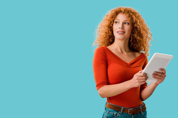 pensive woman with red hair using digital tablet isolated on blue