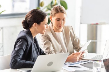 two smiling businesswomen in formal wear at table in office clipart