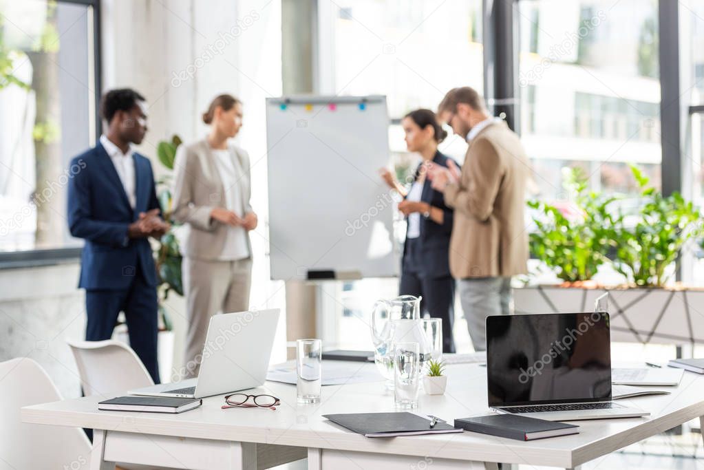 selective focus of four multiethnic businesspeople near flipchart and table with laptops and glasses of water on foreground