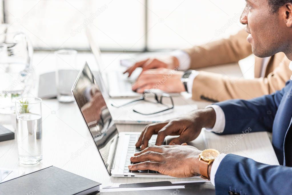 partial view of two multiethnic businessmen using laptops at table in office