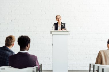 attractive businesswoman standing and smiling during conference in conference hall clipart