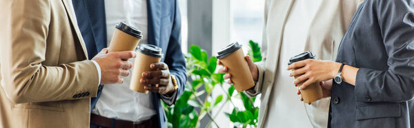 panoramic shot of four multiethnic colleagues holding disposable cups of coffee in office