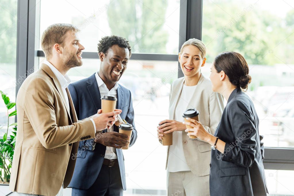 four multiethnic colleagues holding disposable cups of coffee and smiling in office