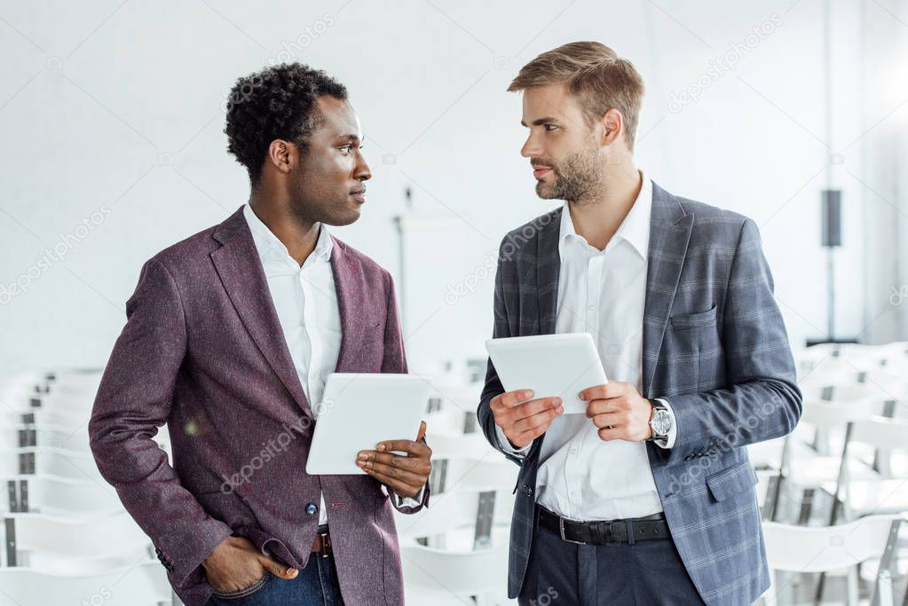 two multiethnic colleagues in formal wear using digital tablets in conference hall