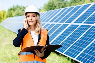 hardhat ve güvenlik yeleği klasörü tutan ve akıllı telefon üzerinde konuşurken çekici iş kadını 