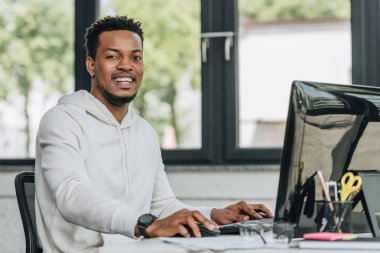 ofiste bilgisayar üzerinde çalışırken kamera bakarak neşeli afrikalı-amerikalı programcı