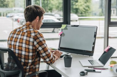 back view of young programmer working on computer while sitting at workplace near window clipart