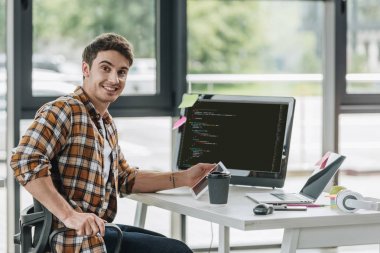 cheerful programmer smiling at camera while sitting near computer monitor with script on screen clipart