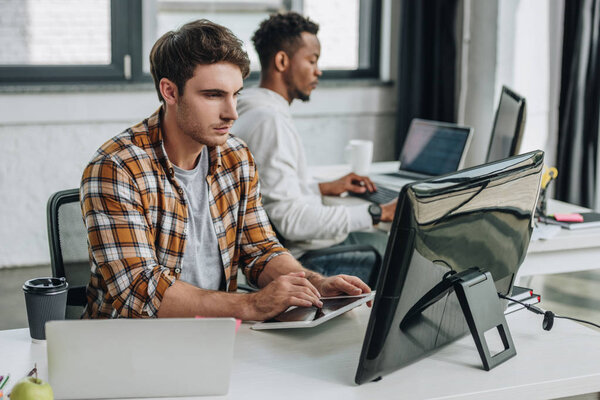 serious young programmer using digital tablet while working in office near african american colleague