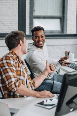 neşeli afro-amerikan programcı akıllı telefon tutan ve başparmak gösteren meslektaşının yanında otururken kamera bakıyor