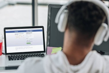 Kiev, Ukrayna - 29 Temmuz 2019: ekranda Facebook web sitesi ile dizüstü bilgisayar yakınında oturan kulaklık afro-amerikan programcı geri görünümü