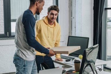 genç afrikalı-Amerikalı programcı gülümseyen meslektaşı nın yanında dururken pizza kutusu tutan