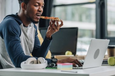 young african american programmer eating pizza while working in office clipart