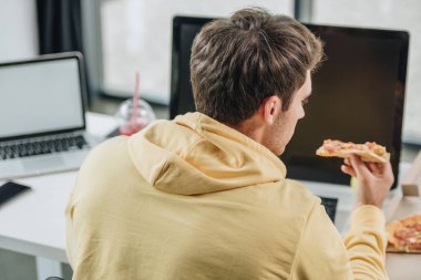 back view of young programmer eating pizza while sitting at workplace in office clipart