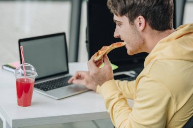 young programmer eating pizza while sitting at workplace in office clipart