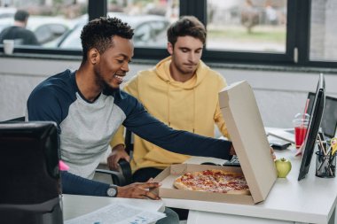 smiling african american programmer opening pizza box while sitting near colleague in office clipart