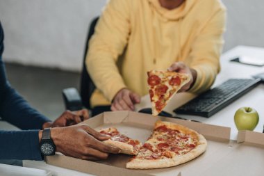 cropped view of two multicultural programmers eating pizza in office clipart