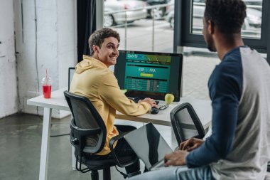 Kiev, Ukrayna - 29 Temmuz 2019: ekranda Sportsbet web sitesi ile bilgisayar kullanan gülümseyen programcı onun Afrikalı Amerikalı meslektaşı dizüstü bilgisayar ile masada otururken