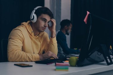 selective focus of thoughtful programmer in headphones working near african american colleague clipart