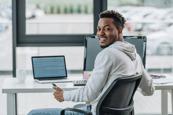 happy african american programmer smiling at camera while sitting at workplace in office