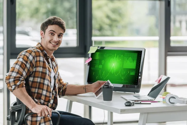 화면에 그래프와 차트와 컴퓨터 모니터 근처에 앉아있는 카메라에 쾌활한 프로그래머 — 스톡 사진