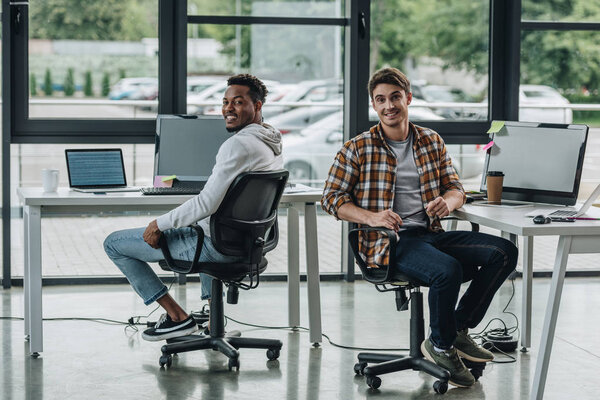 young multicultural programmers smiling at camera while sitting in office chairs