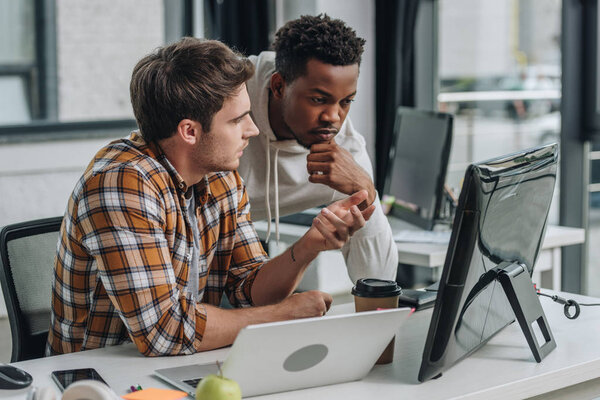 serious programmer gesturing while looking at computer monitor together with african american colleague