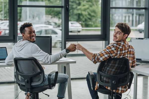 two cheerful multicultural programmers looking at camera while doing fist bump