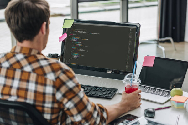 back view of young programmer holding glass of juice while working on computer in office