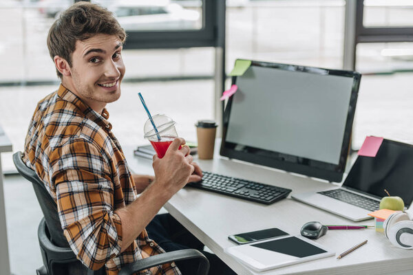 young programmer smiling at camera and holding glass of juice while sitting at workplace 