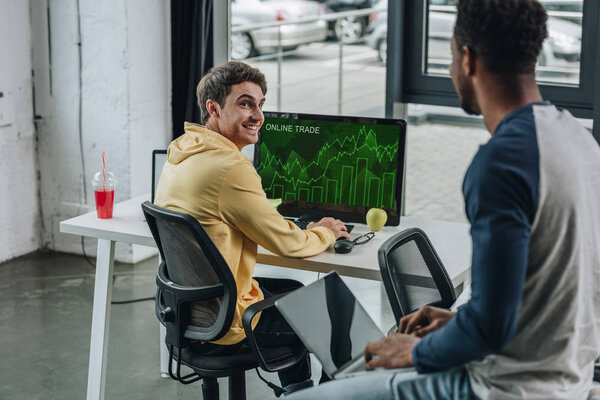 cheerful programmer sitting at computer monitor with online trade on screen and looking at african american colleague