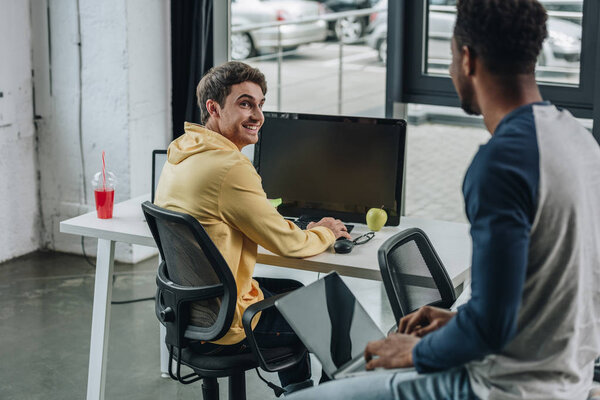 cheerful programmer looking at african american colleague sitting on desk in office